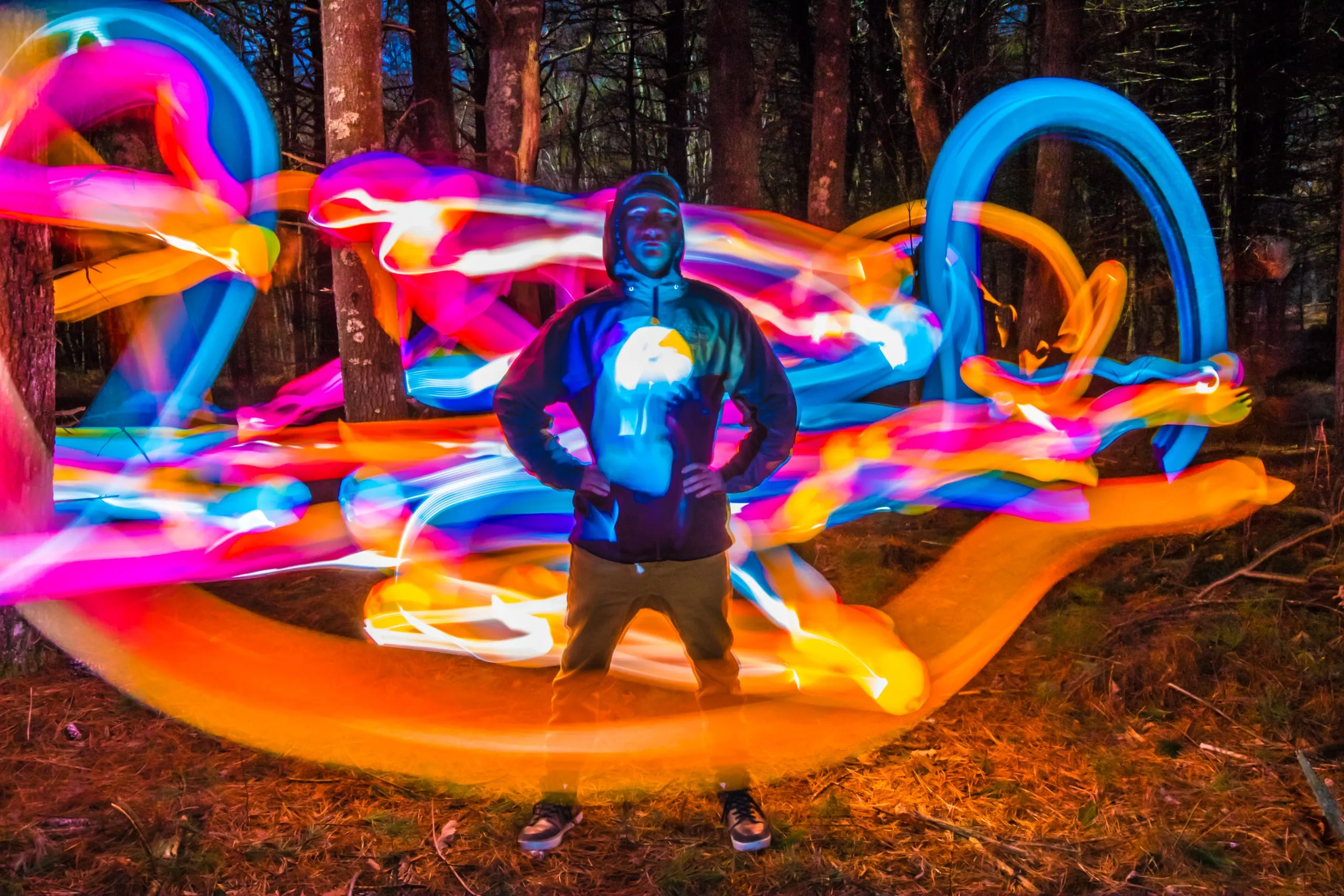 Person standing in swirling multicolored light-painting trails in a forest