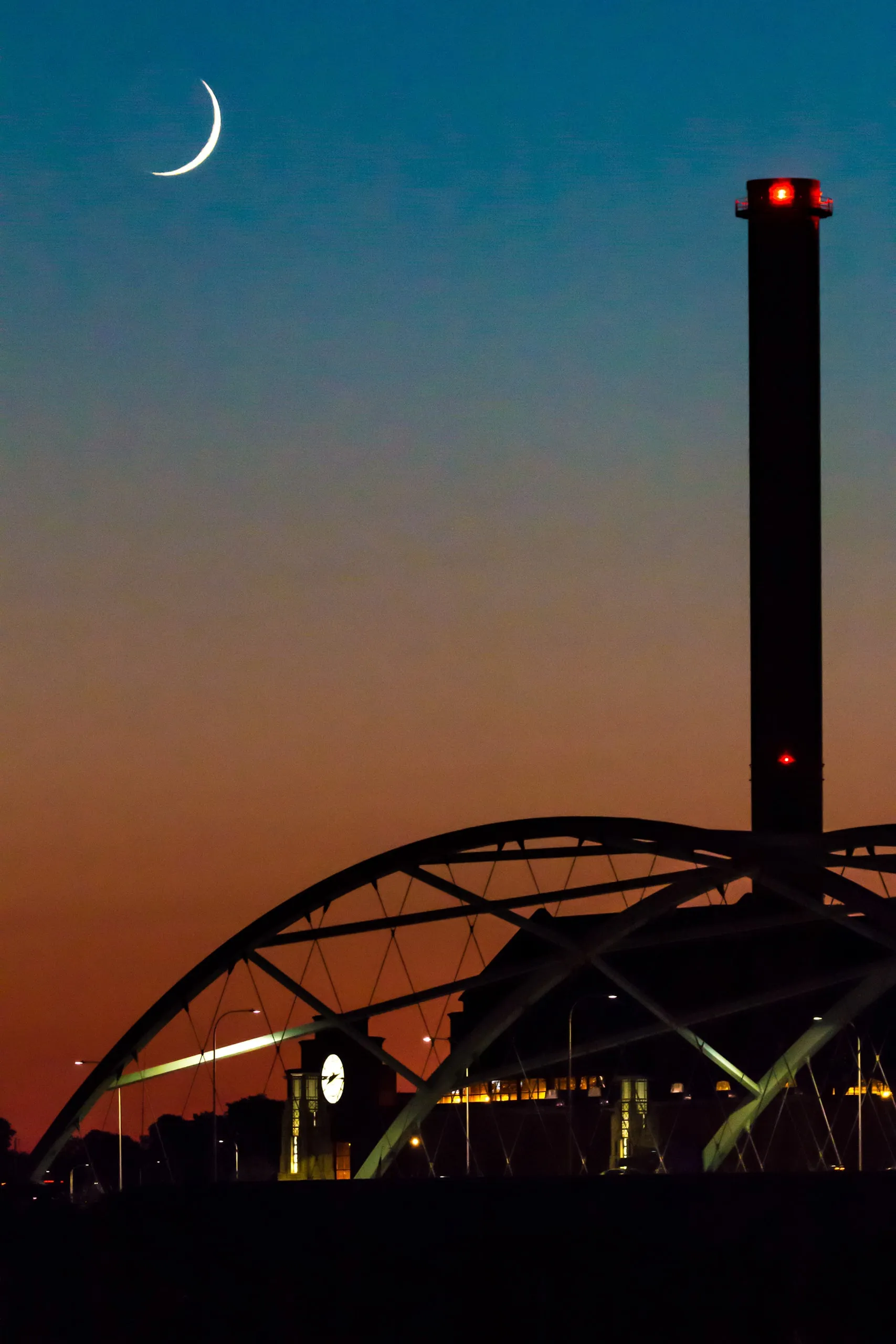 Crescent moon above a smokestack and arched bridge at dusk