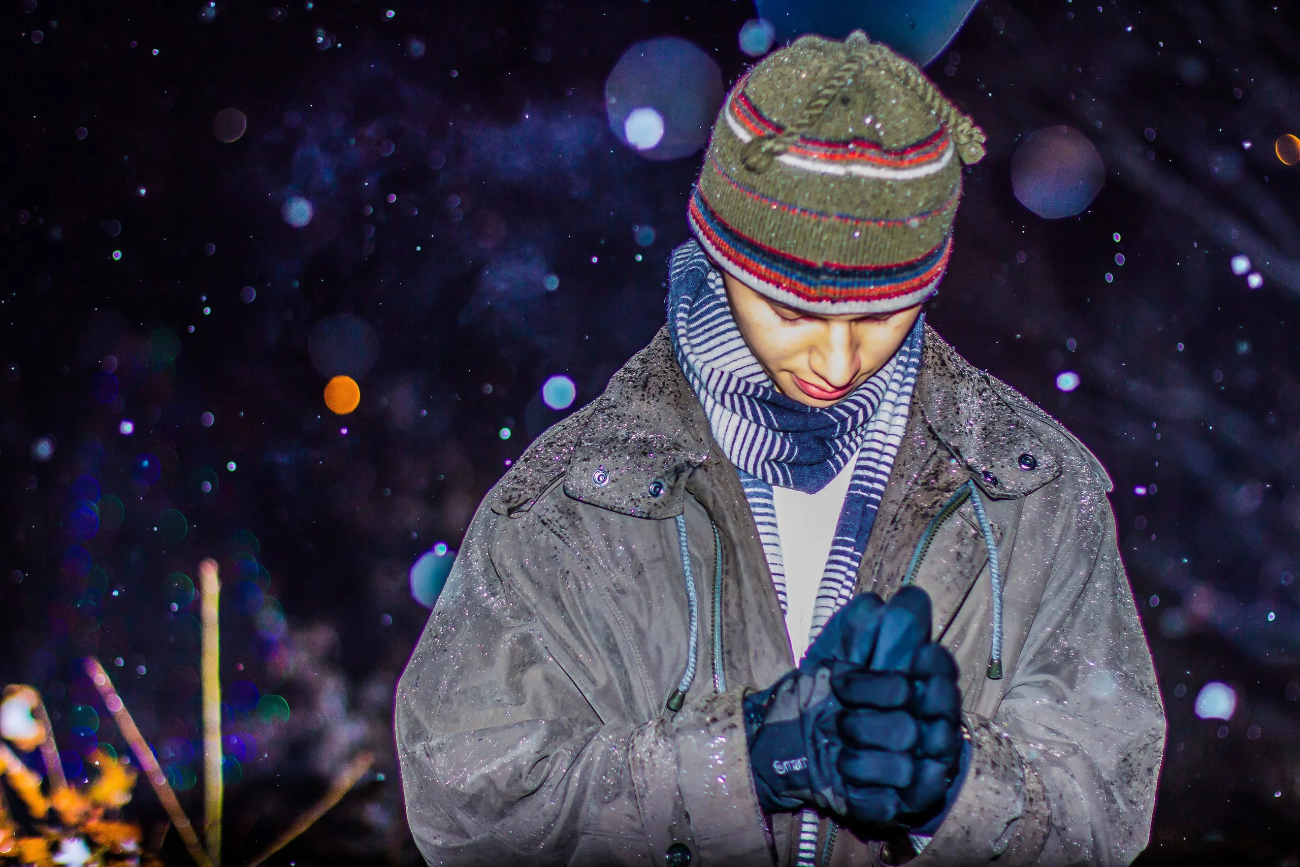 Person in a knit hat and winter coat with hands clasped as snow falls at night