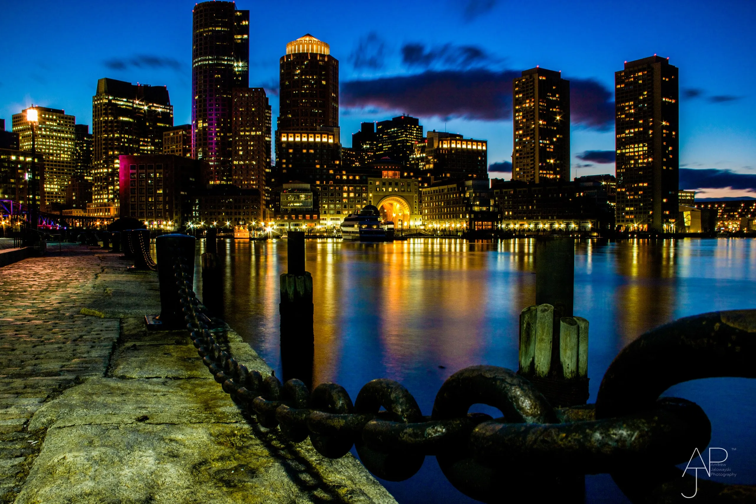 City skyline at night across the harbor, with a chain railing in front